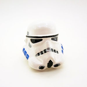 May include: A white plastic figurine of a Star Wars Stormtrooper helmet with blue and black details.