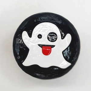 May include: A circular black button featuring a white ghost character with one solid black eye and one circular black eye. The ghost has a wide, open mouth with a bright red tongue sticking out, centered against the glossy black background.