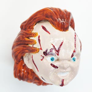 Peut inclure: Sculpture peinte d'une tête de poupée avec des cheveux roux et des yeux bleus. Le visage de la poupée est peint avec des coupures rouges et une expression sinistre. La sculpture est sur fond blanc.