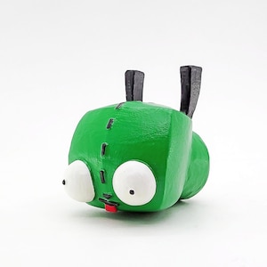 May include: Green and black cartoon character with large white eyes and a red tongue sticking out. The character is made of clay and has a stitched seam down the front.