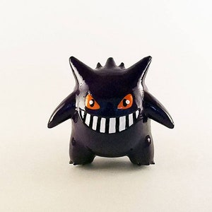 Peut inclure: Une figurine peinte à la main de Gengar, un Pokémon de type Spectre, avec des yeux orange et un large sourire, en noir et blanc.