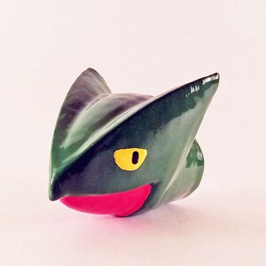 May include: A green ceramic cat-like figurine with a yellow eye and a red mouth. The figurine has pointed ears and a smooth, glossy finish. The text "BossKeyDecor" is written in a teal color at the bottom of the image.