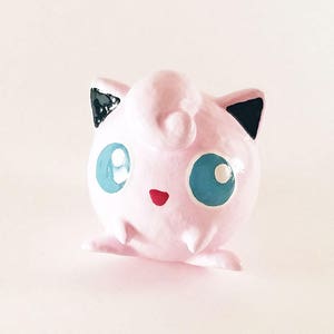 Puede incluir: Una figura redonda rosa con ojos azules grandes, una boca roja pequeña y detalles negros en las orejas. La figura tiene un remolino en la frente y pies pequeños. El texto "BossKeyDecor" está escrito en guión turquesa en la parte inferior.