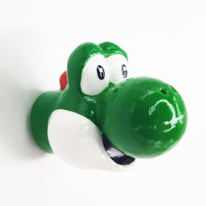 May include: Green Yoshi head figurine with white eyes and a white mouth. The figurine is made of a smooth, glossy material.