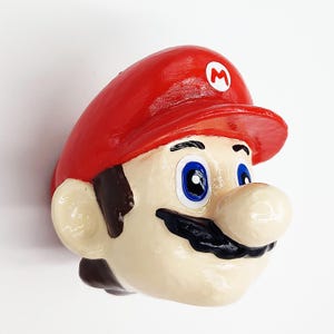 May include: A red and white Mario character figurine with a blue hat and blue eyes. The figurine is made of plastic and has a detailed face.