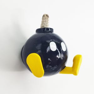May include: A black bomb-shaped wall hook with yellow arms and white eyes. The hook is made of ceramic and has a rope fuse.