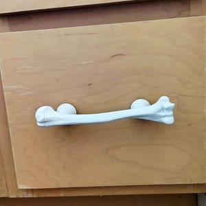 May include: A white drawer pull shaped like a human femur bone, featuring a textured, realistic bone structure with rounded ends. The handle is mounted horizontally onto a light-colored wooden cabinet drawer front with two circular attachment points.