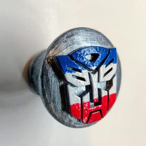 May include: A gray, round cabinet knob featuring the Transformers logo in red, white, and blue. The logo is a stylized face with geometric shapes and is centered on the knob's surface.