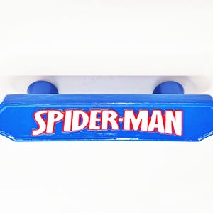 Op de afbeelding: Een blauwe ladeknop met een glanzende afwerking voorzien van de tekst SPIDER-MAN in witte letters met een rode omlijning. De handgreep heeft een rechthoekige vorm met schuine uiteinden en twee ronde bevestigingspunten zichtbaar aan de achterzijde.