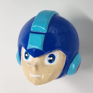 May include: A blue and light blue plastic figurine of a robot head with a white face and blue eyes.