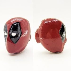 May include: A red and black Deadpool head figurine. The head is made of ceramic and has a glossy finish. The eyes are white and the mask is black with a red outline.
