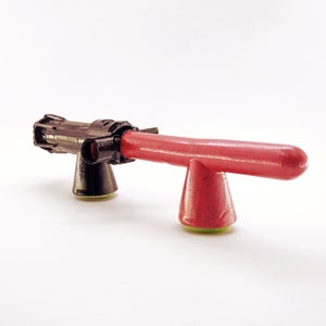 Peut inclure: Un jouet pistolet noir et rouge avec un canon pointé vers la droite. Le pistolet est sur une surface blanche.