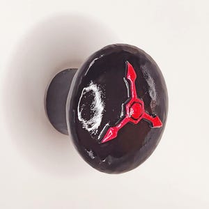 May include: A round, black cabinet knob with a glossy finish. The front surface features a stylized, three-pointed red graphic resembling clock hands or a geometric symbol, along with a small, textured white circular mark on the left side.