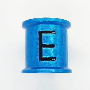 May include: A cylindrical, blue object with a black letter "E" in the center. The object has a textured surface and a rounded top and bottom. The background is plain white.