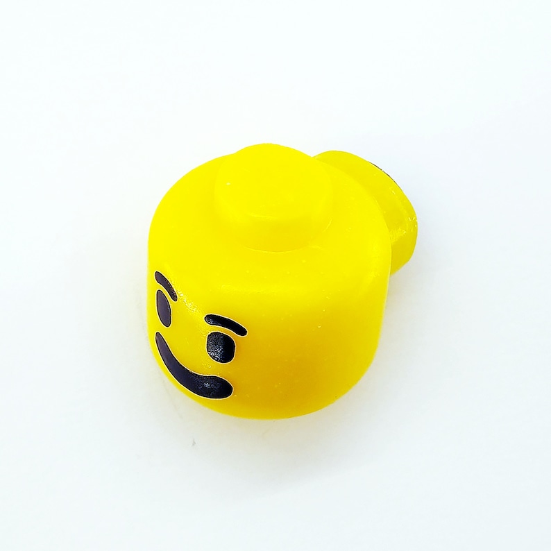 May include: Yellow plastic Lego head with a black smiling face and a single eyebrow raised.