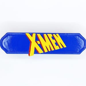 Puede incluir: Un pin rectangular azul con bordes biselados que presenta el texto amarillo X-MEN en una fuente negrita y estilizada. Las letras están en relieve y tienen un ligero contorno rojo, creando un efecto tridimensional sobre la superficie azul lisa y brillante.