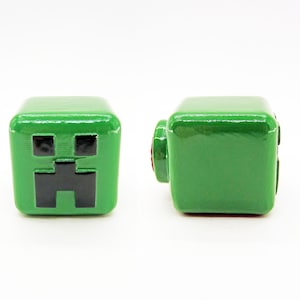 May include: Two green Minecraft Creeper block figures. One figure has a black Creeper face on the front. The other figure is plain green.