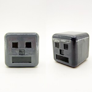 May include: Two gray cube-shaped objects with a simple face design. Each cube has two black eyes, a black nose, and a black mouth.