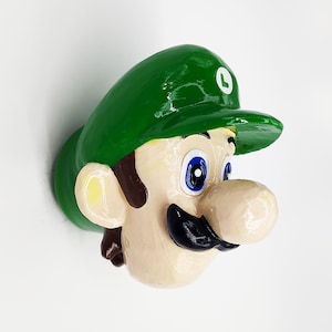 May include: A green and white figurine of a character with a green cap, brown hair, a brown mustache, and a large nose. The character has blue eyes and a white circle on the cap.