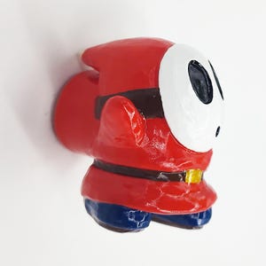 May include: A red and white figurine of a character from the video game Super Mario Bros. The character is wearing a red hooded cloak and a white mask with black eyes. The figurine is standing on a white surface.