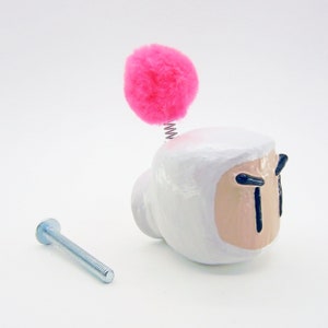 May include: White ceramic door knob with a pink pom-pom on top. The knob has a black and white face with a frowning expression. A silver screw is included.