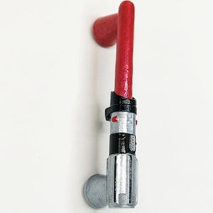 May include: A vertical cabinet pull shaped like a red lightsaber with a silver and black hilt. The handle features a textured red blade extending from a detailed metallic-colored grip with small red accents and a silver mounting base.