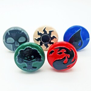 May include: Five ceramic round tokens with different symbols. The symbols are a skull, a sun with rays, a tree, a drop of water, and a flame. The tokens are in different colors: gray, beige, green, blue, and red.