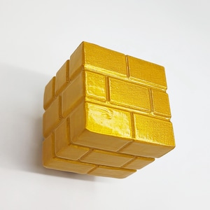 May include: A gold-painted, brick-patterned cube, likely a decorative item or puzzle. The cube's surface has a textured appearance, with each brick clearly defined. The object is set against a plain white background, highlighting its form and color.