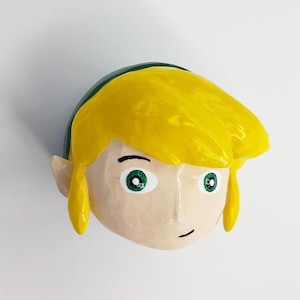 May include: A 3D printed figurine of Link from the Legend of Zelda video game series. The figurine is painted with green, yellow, and white paint. Link has a green cap, yellow hair, and green eyes.