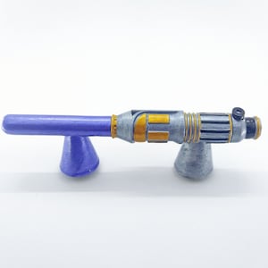 May include: A silver and gold-coloured toy lightsaber with a purple handle and a black tip. The lightsaber is on a purple stand.