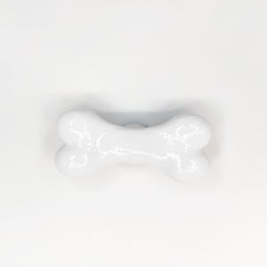 May include: A white ceramic drawer knob shaped like a dog bone. The knob features a smooth, glossy finish with rounded ends and a central indentation, designed for attachment to furniture surfaces with a single mounting point on the back.