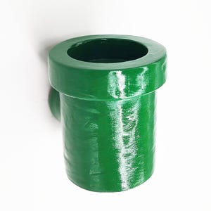 May include: A green plastic pipe-shaped holder for a toothbrush or other small items. It has a smooth, glossy finish and a rounded top.