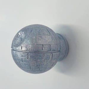 May include: A silver-coloured Death Star-shaped wall hook. The hook is designed to look like the Death Star from the Star Wars films. It has a textured surface and a small hole on the side for hanging.