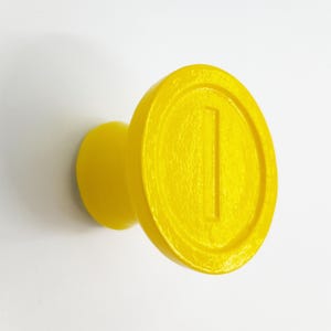 May include: A yellow plastic wall hook shaped like a Super Mario coin. The coin has a raised line down the center.