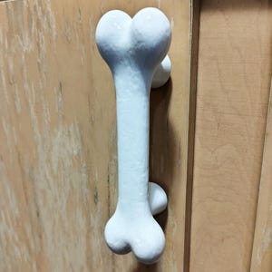 May include: A white cabinet handle shaped like a bone. The handle features a smooth, matte finish and is mounted vertically onto a light-colored wooden cabinet door. The design mimics the anatomical structure of a long bone with rounded ends.