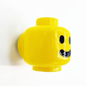 May include: A yellow plastic Lego head with a black smiling face. The head is a wall-mounted decoration.