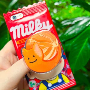 May include: A red smartphone case featuring the white text Milky and Japanese characters, with an attached orange circular phone grip displaying a smiling cat-like character with a stylized orange slice design on its back.