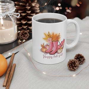 May include: White ceramic mug with a fall design. The design features pink wellington boots, pumpkins, and autumn leaves. The text "Happy fall y'all" is written in a script font. The mug is filled with a dark liquid.