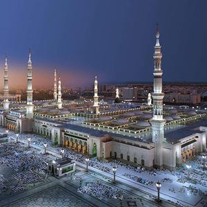 May include: An aerial view of a large mosque complex at dusk. The mosque features numerous tall minarets and a large central courtyard. The architecture is primarily white and beige, with many lights illuminating the structure and surrounding area.