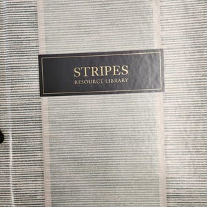 May include: A wallpaper sample featuring a striped design in grey and white tones. The text "STRIPES RESOURCE LIBRARY" is on a black label with gold edging. The brand name "YORK WALLCOVERINGS" is printed in black at the bottom.