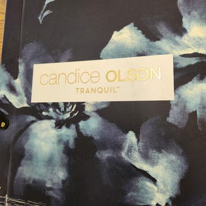 Puede incluir: Un muestrario de tela negro con una etiqueta blanca que dice "candice OLSON TRANQUIL" en letras doradas. La tela tiene un patrón floral azul y blanco.