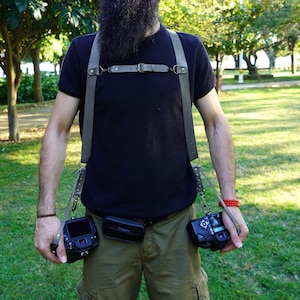 May include: A man with dreadlocks and a beard is wearing a black t-shirt and khaki cargo trousers. He is wearing a leather camera strap with two cameras attached. The cameras are black and have a silver lens.