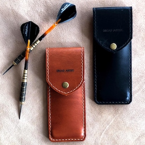 May include: Two darts with black and orange flights next to two leather dart cases. One case is tan, the other is black. The tan case has the text "BREAD ARTIST" stitched on it. The cases have a snap closure.