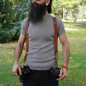 May include: A man with a long beard and dreadlocks is wearing a grey t-shirt and brown leather camera straps. He is holding two black cameras in front of him.