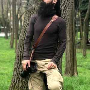 May include: A man with a long beard and dreadlocks is wearing a brown turtleneck sweater and khaki pants. He is holding a camera with a brown leather camera strap. He is standing in a forest with trees in the background.