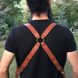 Puede incluir: Correas de cuero marrón con herrajes de latón forman un arnés en forma de X. Las correas son ajustables y tienen un anillo en el centro.