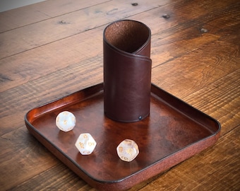 May include: A brown leather dice rolling tray with a matching leather dice tower. Three white dice with gold numbers are on the tray.