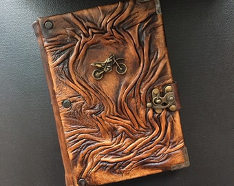 Pode incluir: Um diário de couro marrom com um fecho de metal decorativo e um pequeno design de motocicleta de metal na capa.