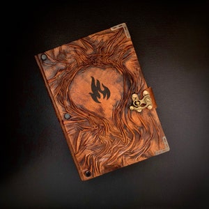Peut inclure: Un journal relié en cuir marron avec un motif gaufré décoratif et un fermoir en métal. Le journal a un symbole de flamme noire sur la couverture.
