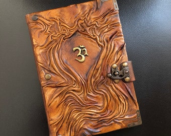 Könnte beinhalten: Ein braunes Lederjournal mit einem dekorativen Verschluss und einem goldenen Om-Symbol auf dem Einband.